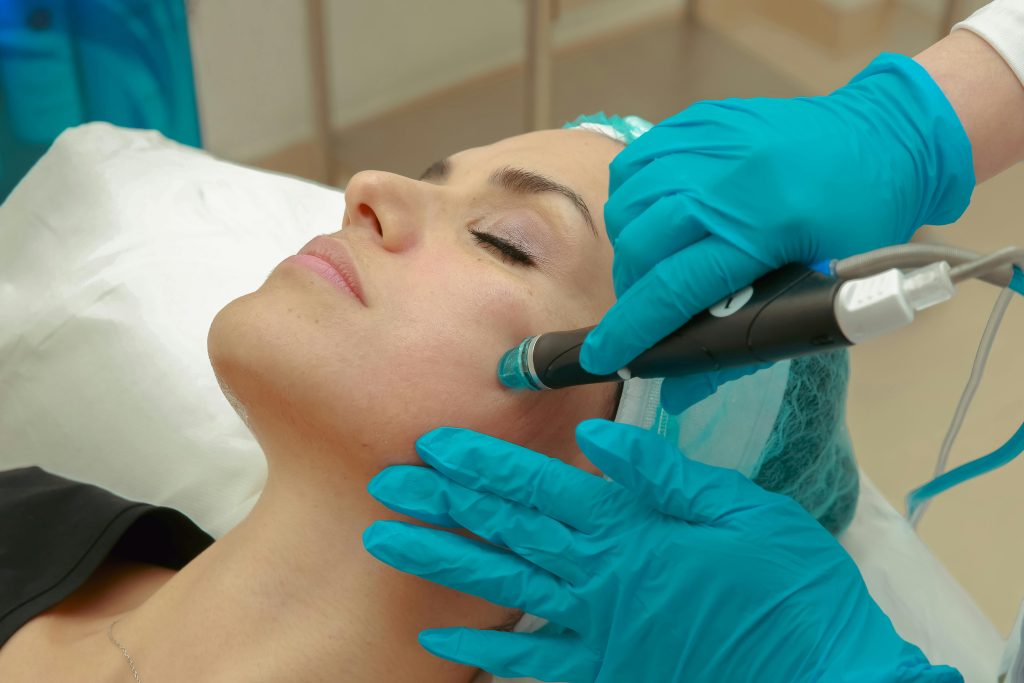 A woman receiving a facial skincare treatment with a specialized device wearing protective gloves.